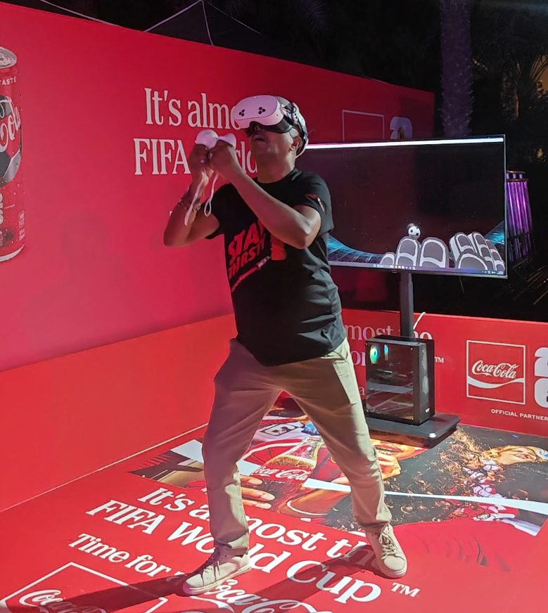A player wearing a VR headset defending a goal at a Coca-Cola FIFA World Cup activation, with the game view shown on a screen beside them.